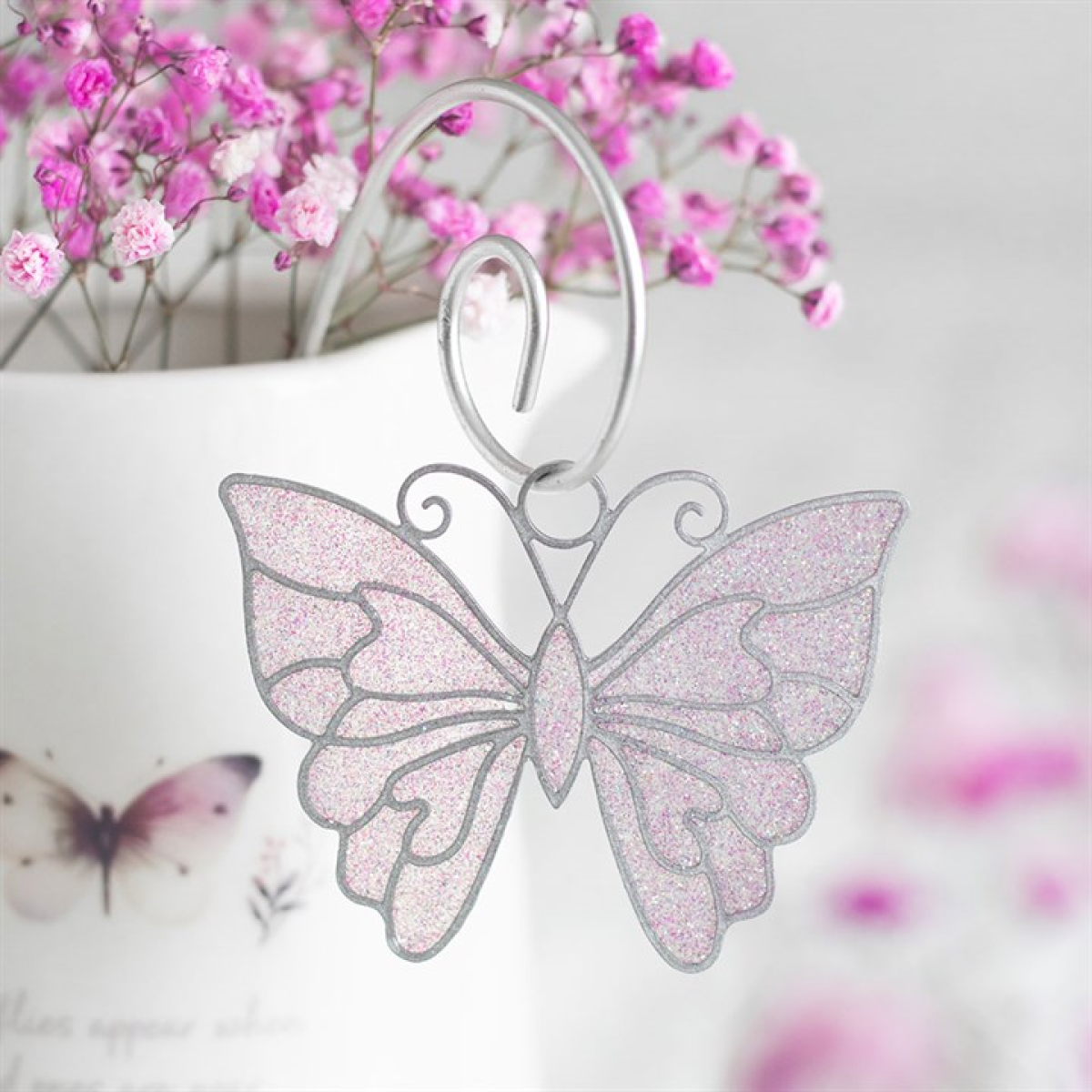 Butterfly-shaped ornament with pink glitter on a white background with blurred pink flowers.